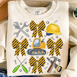 May include: Cream-colored sweatshirt featuring a construction-themed graphic. The design includes a yellow and black striped bow, a yellow hard hat, a small excavator, wrenches, screws, and a hammer and saw.