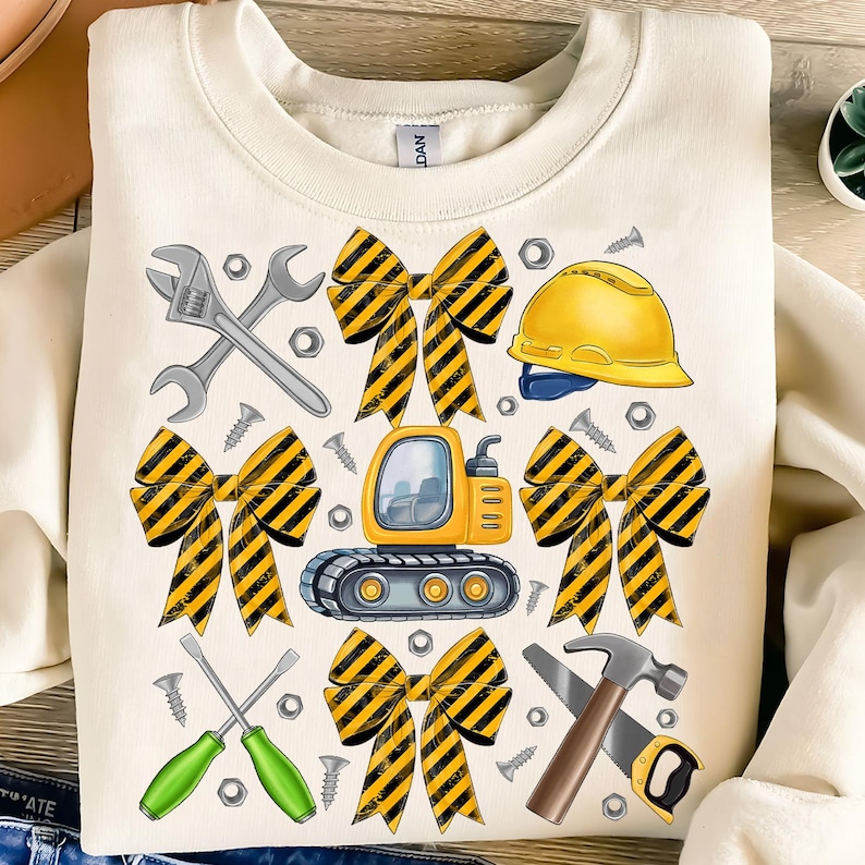 May include: Cream-colored sweatshirt featuring a construction-themed graphic. The design includes a yellow and black striped bow, a yellow hard hat, a small excavator, wrenches, screws, and a hammer and saw.