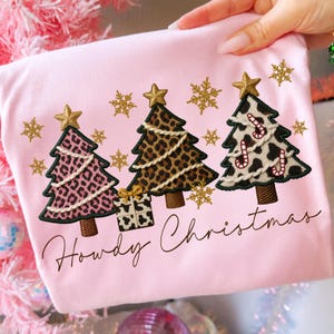 Pu&ograve; includere: T-shirt rosa chiaro con alberi di Natale ricamati con motivi animalier. Gli alberi hanno stelle dorate e fiocchi di neve. Il testo "Howdy Christmas" &egrave; scritto in un carattere corsivo.