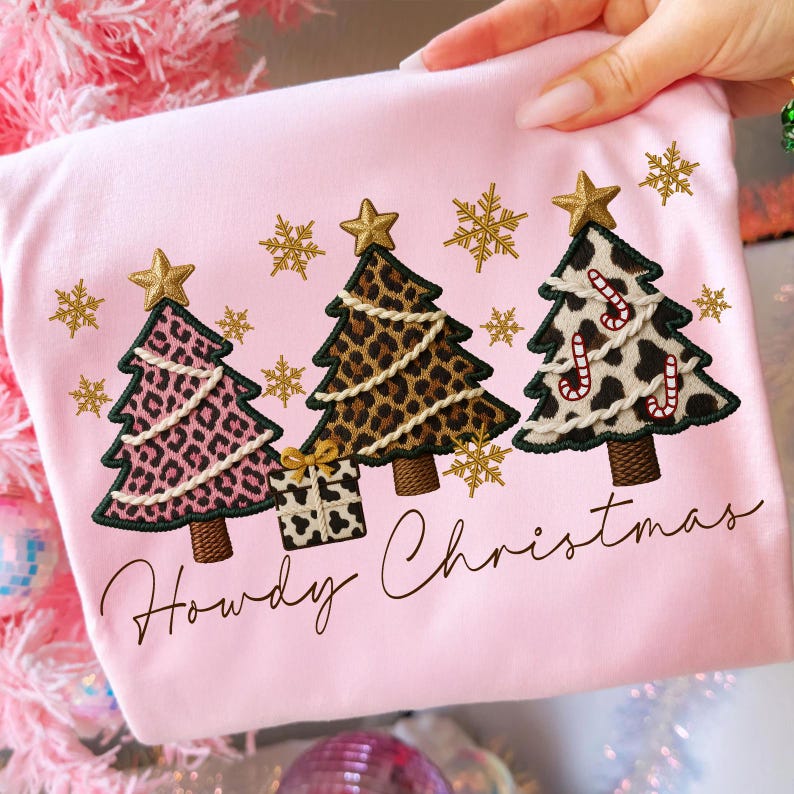 Pu&ograve; includere: T-shirt rosa chiaro con alberi di Natale ricamati con motivi animalier. Gli alberi hanno stelle dorate e fiocchi di neve. Il testo "Howdy Christmas" &egrave; scritto in un carattere corsivo.