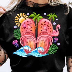 May include: Black t-shirt featuring a vibrant graphic of pink clogs adorned with seashells and starfish. The design includes a sun, palm trees, a flamingo, jellyfish, and ocean waves, creating a summery theme.