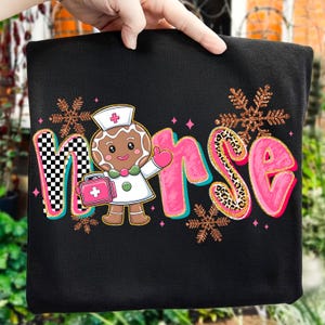 May include: Black sweatshirt with a colorful graphic that says "Nurse". The word "Nurse" is designed with a gingerbread nurse character, pink letters, and snowflake accents. The letter "N" has a black and white checkered pattern.