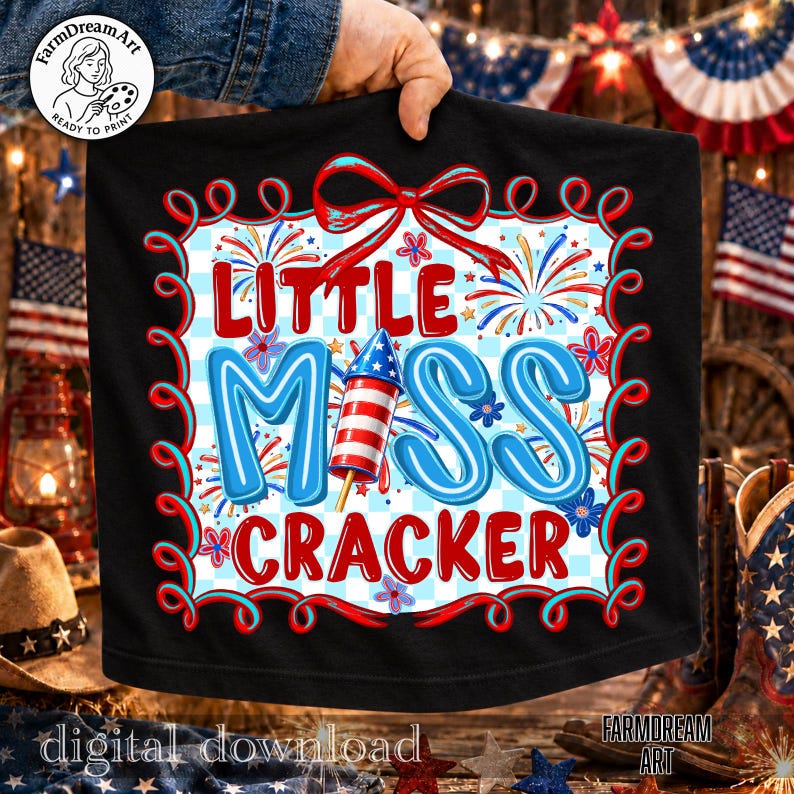 May include: Black t-shirt with a patriotic design. The shirt features the text "Little Miss Cracker" in red and blue, with a rocket and fireworks graphic. The design is framed by a red and blue border.