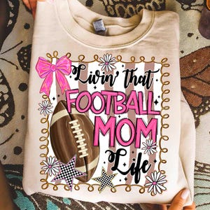May include: A beige sweatshirt with the text "Livin' that FOOTBALL MOM Life" in black and pink. The design includes a football, pink bow, daisies, and checkered stars. The sweatshirt is folded.