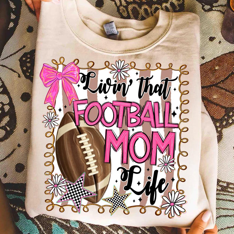 May include: A beige sweatshirt with the text "Livin' that FOOTBALL MOM Life" in black and pink. The design includes a football, pink bow, daisies, and checkered stars. The sweatshirt is folded.