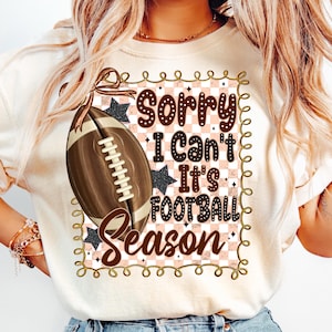 May include: Cream-colored t-shirt with a football graphic and the text "Sorry I Can't It's Football Season." The design includes a brown football, black stars, and a pink and white checkered background.
