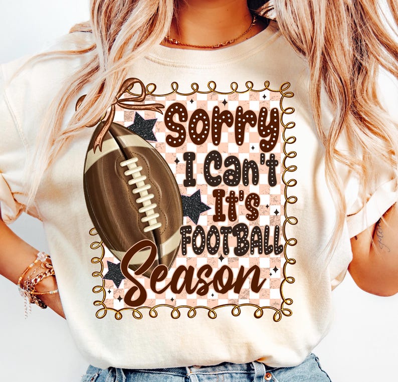 May include: Cream-colored t-shirt with a football graphic and the text "Sorry I Can't It's Football Season." The design includes a brown football, black stars, and a pink and white checkered background.