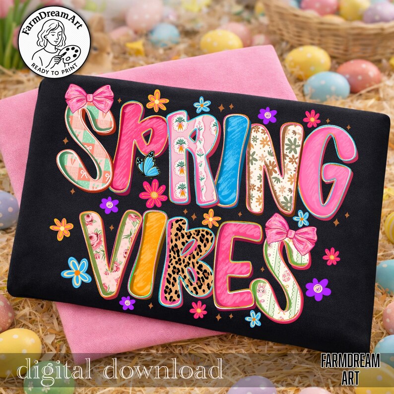 May include: Black t-shirt with the words "Spring Vibes" in colorful, floral, and patterned letters. The design includes butterflies, flowers, and bows. The words are outlined in a bright pink color. The shirt is on a pink surface.