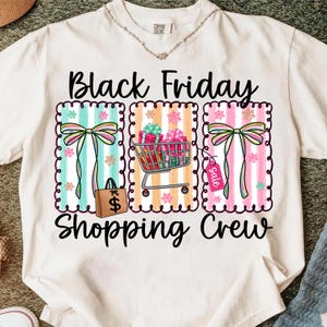 May include: Cream-colored t-shirt with a graphic design featuring the text "Black Friday Shopping Crew." The design includes colorful striped panels with bows, a shopping cart filled with gifts, and a shopping bag.