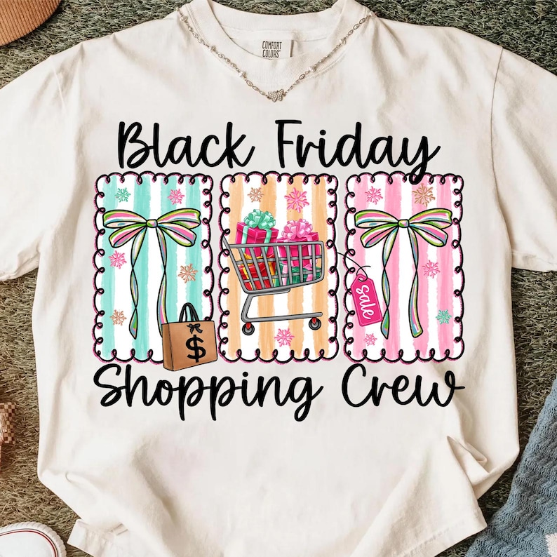 May include: Cream-colored t-shirt with a graphic design featuring the text "Black Friday Shopping Crew." The design includes colorful striped panels with bows, a shopping cart filled with gifts, and a shopping bag.