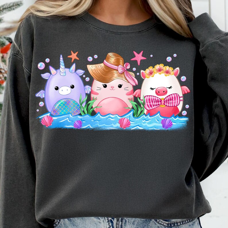 K&ouml;nnte beinhalten: Ein dunkelgrauer Pullover mit einem Grafikdruck von drei Comicfiguren. Die Figuren sind ein lilafarbenes Einhorn, eine rosa Katze mit Hut und ein wei&szlig;es Schwein mit Fliege. Alle drei Figuren befinden sich in einem Wasserbecken mit Muscheln und Seegras.
