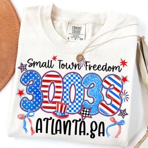 May include: Cream-colored t-shirt with the text "Small Town Freedom" and "30033 ATLANTA.GA" in patriotic colors. The design includes stars, stripes, and a top hat. A gold necklace rests on the shirt.