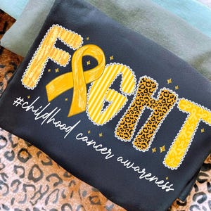 Puede incluir: Camiseta negra con la palabra "FIGHT" en amarillo, estampado de leopardo y patrones de rayas, con una cinta amarilla. El texto "#childhood cancer awareness" está escrito debajo de la palabra "FIGHT".