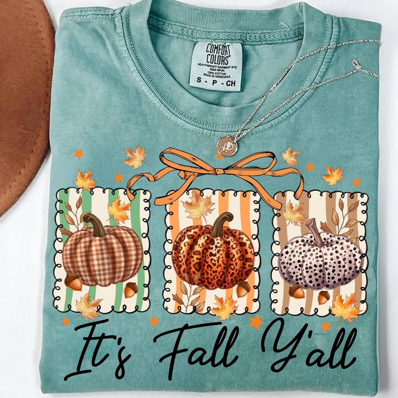 May include: A teal Comfort Colors t-shirt featuring three pumpkin illustrations with fall leaves and the text "It's Fall Y'all". The pumpkins have plaid, leopard, and polka dot patterns. A silver necklace with a pendant is draped across the shirt.