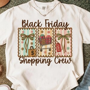 May include: White t-shirt with a graphic design featuring the text "Black Friday Shopping Crew." The design includes three rectangular frames with bows, shopping bags, a shopping cart, and a sale tag. The shirt is made of a soft material.