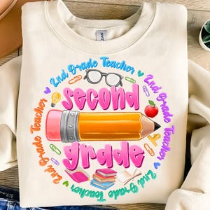 May include: Cream-colored sweatshirt with a colorful graphic design for a second-grade teacher. The design includes a large pencil, the words "Second Grade" in pink, and other school-related icons like glasses, an apple, and books.