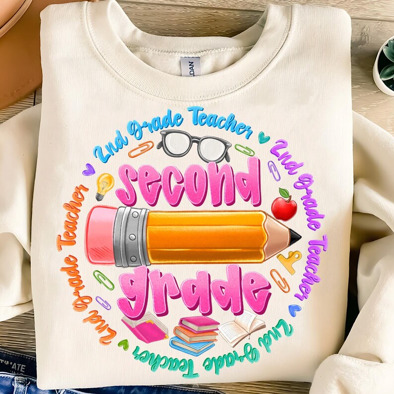 May include: Cream-colored sweatshirt with a colorful graphic design for a second-grade teacher. The design includes a large pencil, the words "Second Grade" in pink, and other school-related icons like glasses, an apple, and books.