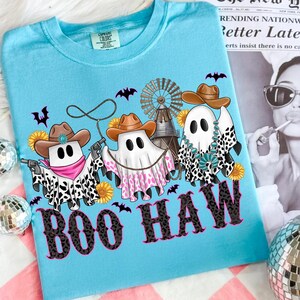 May include: Light blue t-shirt with a Halloween-themed graphic. The design features three ghost figures wearing cowboy hats and bandanas, holding guns, and surrounded by bats, sunflowers, and a windmill. The text "BOO HAW" is printed in a leopard print font.