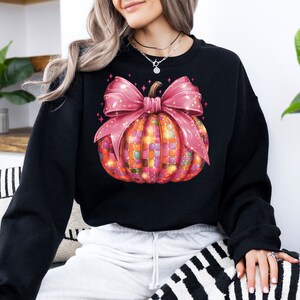 May include: Black sweatshirt featuring a colorful pumpkin design with a pink bow. The pumpkin is made of small, colorful squares, and the bow has glitter accents. The design is centered on the front of the sweatshirt, creating a festive fall look.