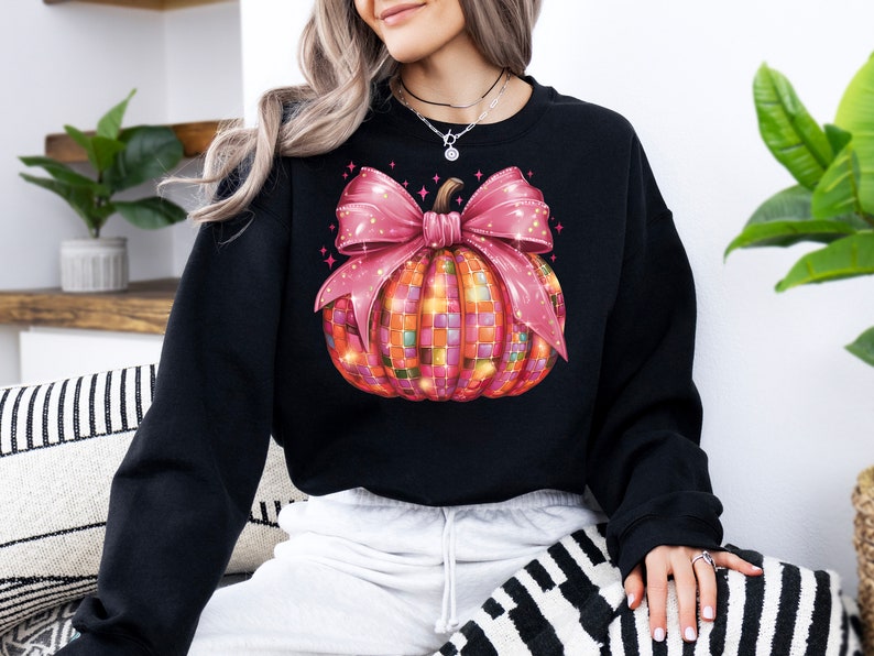 May include: Black sweatshirt featuring a colorful pumpkin design with a pink bow. The pumpkin is made of small, colorful squares, and the bow has glitter accents. The design is centered on the front of the sweatshirt, creating a festive fall look.