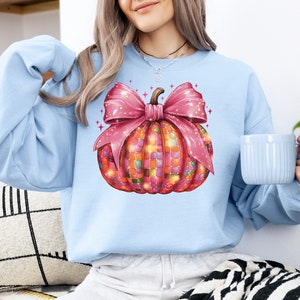 May include: Light blue sweatshirt featuring a colorful pumpkin design. The pumpkin is decorated with a mosaic pattern and a large pink bow. The design is festive and suitable for fall or Halloween.