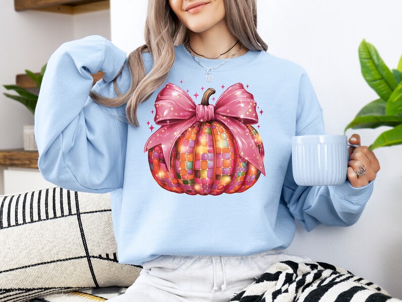 May include: Light blue sweatshirt featuring a colorful pumpkin design. The pumpkin is decorated with a mosaic pattern and a large pink bow. The design is festive and suitable for fall or Halloween.