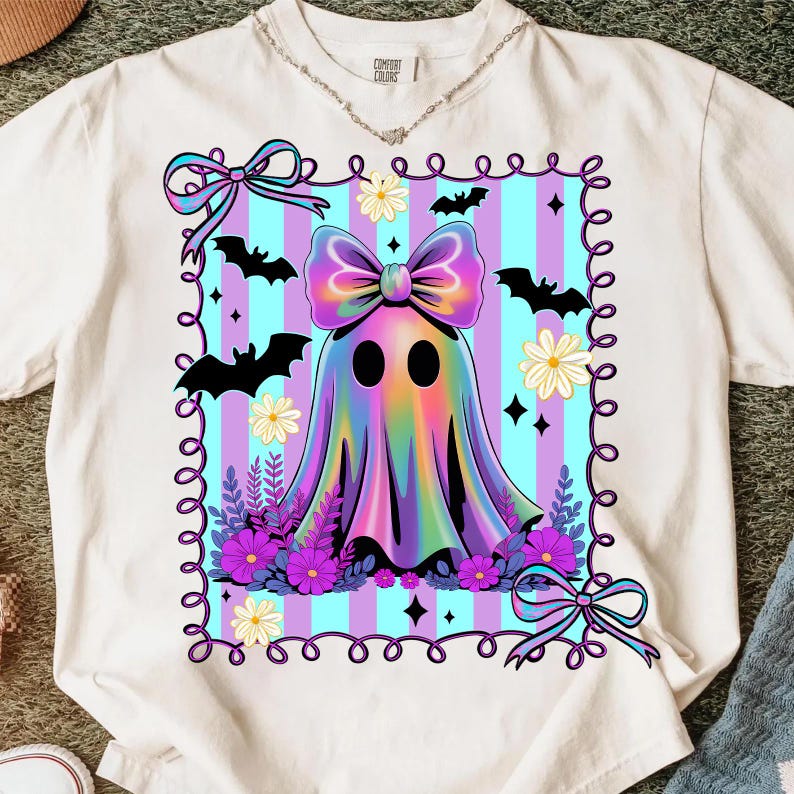 May include: Cream-colored t-shirt featuring a colorful ghost design. The ghost has a large bow and is surrounded by bats, flowers, and bows. The background has blue and purple stripes, perfect for Halloween.