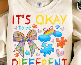 May include: A white sweatshirt with the text "It's okay to be different" in colorful crayon-like lettering. The design includes a rainbow, a bow, and puzzle pieces.