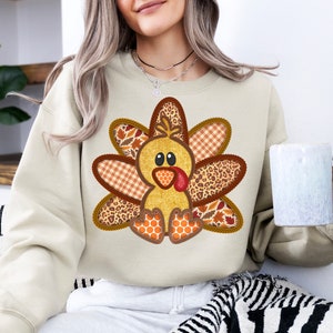 May include: A beige sweatshirt featuring a patchwork turkey design. The turkey is made from brown, orange and white fabric with a black and white leopard print. The turkey has a red beak and wattle.