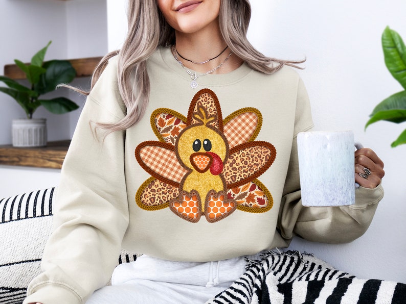 May include: A beige sweatshirt featuring a patchwork turkey design. The turkey is made from brown, orange and white fabric with a black and white leopard print. The turkey has a red beak and wattle.