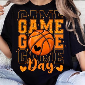 Peut inclure: T-shirt noir avec un texte en paillettes orange qui dit "Game Game Game Day" et un grand ballon de basket orange avec un c&oelig;ur au centre.