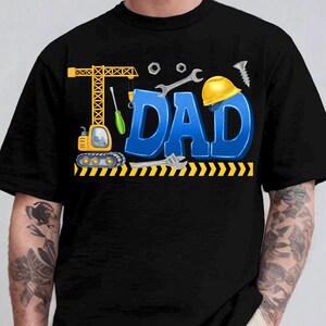 May include: Black t-shirt with a colorful graphic design. The design features the word "DAD" in blue 3D letters, construction tools, a yellow hard hat, and a crane. The shirt is a crew neck.