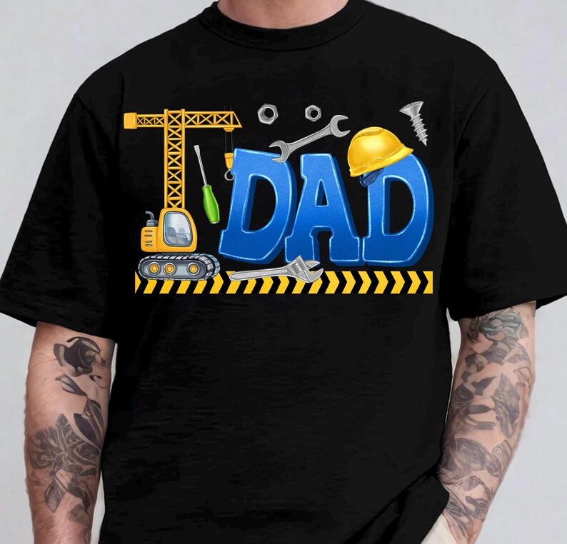 May include: Black t-shirt with a colorful graphic design. The design features the word "DAD" in blue 3D letters, construction tools, a yellow hard hat, and a crane. The shirt is a crew neck.