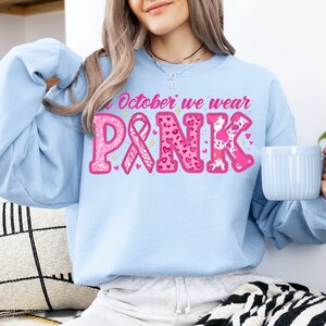 May include: A light blue sweatshirt with the text "In October we wear PINK" in pink letters with glitter and pink ribbons. The letters are decorated with pink hearts, leopard print, and zebra print.