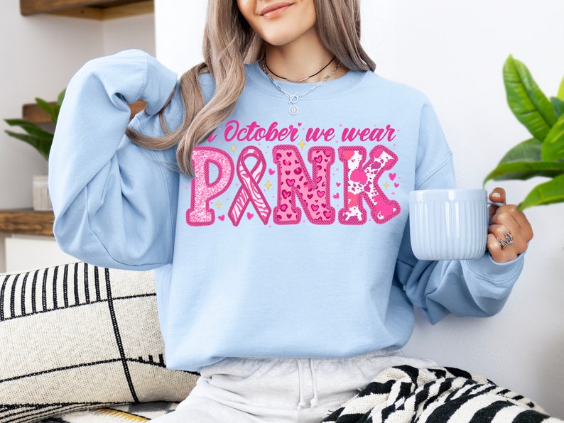 May include: A light blue sweatshirt with the text "In October we wear PINK" in pink letters with glitter and pink ribbons. The letters are decorated with pink hearts, leopard print, and zebra print.