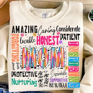 May include: A white sweatshirt with a colorful graphic design that says "MOM" surrounded by words describing a mother's qualities, such as "amazing", "caring", "honest", "patient", "inspiring", "protective", "nurturing", "dependable", "supportive", and "beautiful".