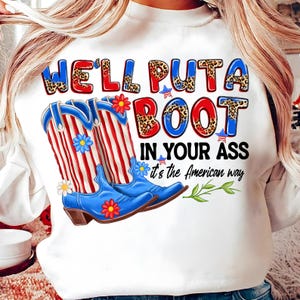 May include: White sweatshirt with a graphic design featuring blue cowboy boots with red and white stripes, and the text "WE'LL PUTA BOOT IN YOUR ASS it's the American way". The boots have floral accents.