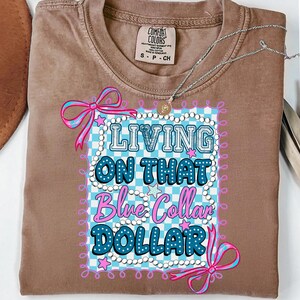 Pu&ograve; includere: Una maglietta marrone Comfort Colors con la scritta "LIVING ON THAT Blue Collar DOLLAR" in un design blu e rosa. La maglietta ha un girocollo e maniche corte. Una collana d'argento con un ciondolo &egrave; sopra la maglietta.