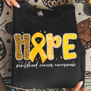 May include: Black sweatshirt with the word "HOPE" in yellow and leopard print. A yellow ribbon is in the center. The text "#childhood cancer awareness" is below the word "HOPE".