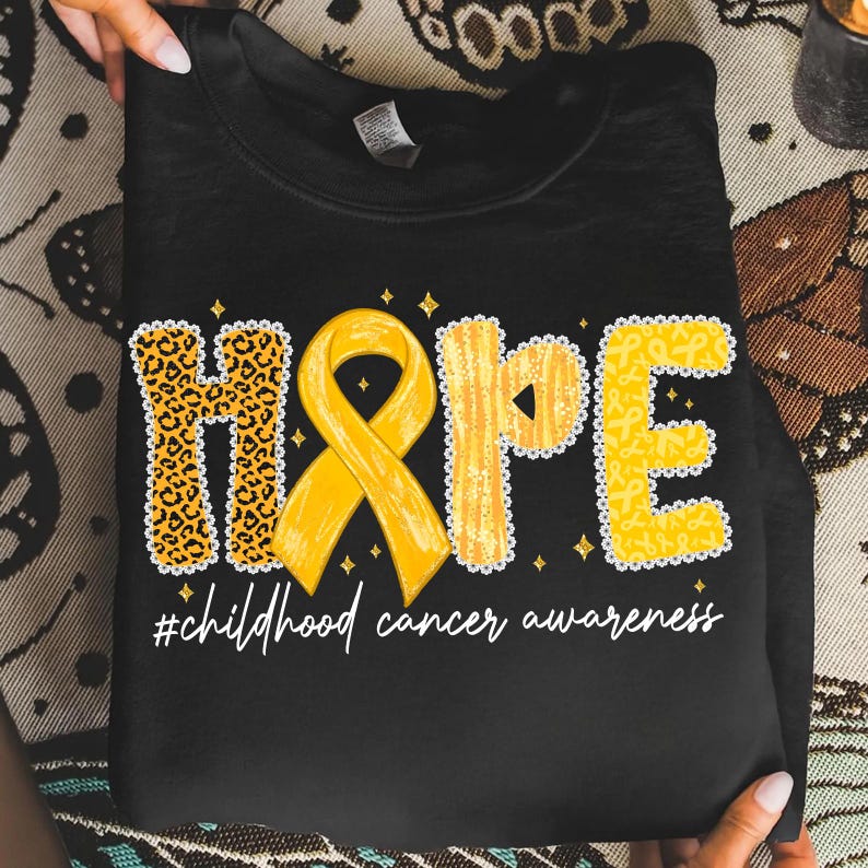 May include: Black sweatshirt with the word "HOPE" in yellow and leopard print. A yellow ribbon is in the center. The text "#childhood cancer awareness" is below the word "HOPE".