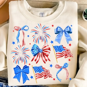 May include: A cream-colored sweatshirt with a festive design featuring red, white, and blue bows, fireworks, and American flags.