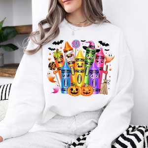 May include: White sweatshirt featuring a colorful Halloween-themed crayon design. The graphic includes smiling crayons dressed as a mummy, witch, and pumpkin, along with bats, ghosts, and jack-o'-lanterns. A fun and festive design for the spooky season.