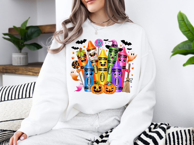 May include: White sweatshirt featuring a colorful Halloween-themed crayon design. The graphic includes smiling crayons dressed as a mummy, witch, and pumpkin, along with bats, ghosts, and jack-o'-lanterns. A fun and festive design for the spooky season.