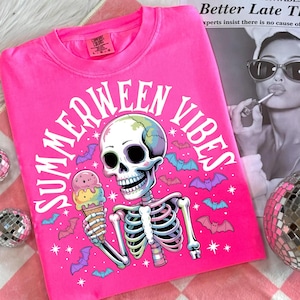 May include: Bright pink t-shirt with the words "SUMMERWEEN VIBES" in a circular design. The graphic features a skeleton holding an ice cream cone, surrounded by bats and stars. The shirt is a Comfort Colors brand.