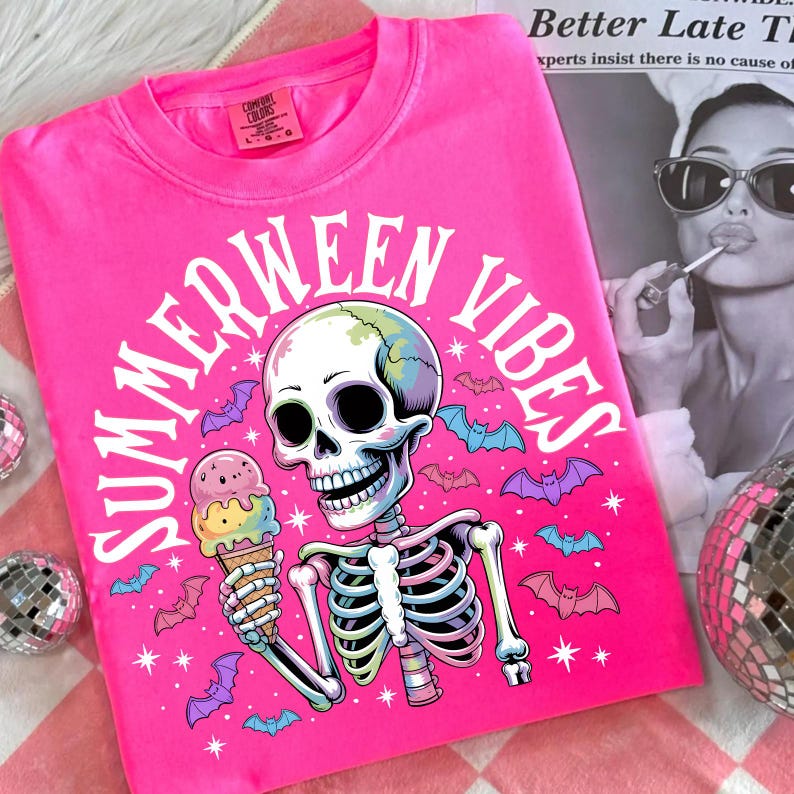 May include: Bright pink t-shirt with the words "SUMMERWEEN VIBES" in a circular design. The graphic features a skeleton holding an ice cream cone, surrounded by bats and stars. The shirt is a Comfort Colors brand.