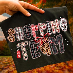 Peut inclure: T-shirt noir avec l'inscription "SHOPPING TEAM" dans un motif patchwork. Les lettres pr&eacute;sentent divers motifs, notamment des imprim&eacute;s l&eacute;opard, des carreaux et des losanges. Un n&oelig;ud rouge et une &eacute;tiquette "soldes" sont &eacute;galement visibles.