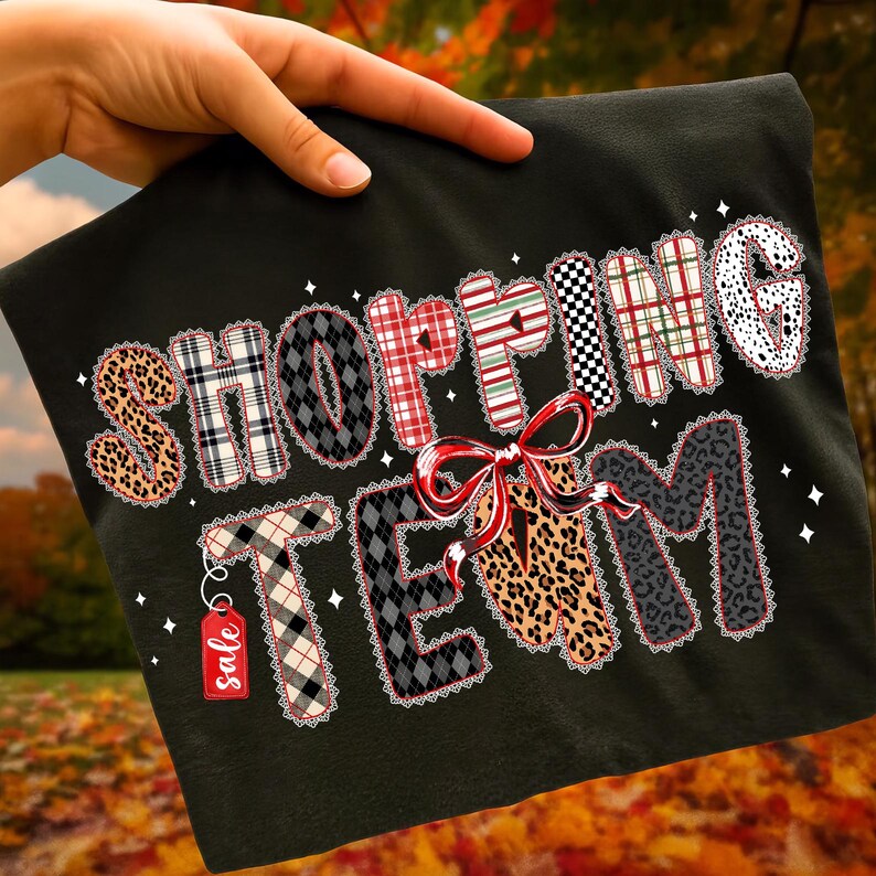 Peut inclure: T-shirt noir avec l'inscription "SHOPPING TEAM" dans un motif patchwork. Les lettres pr&eacute;sentent divers motifs, notamment des imprim&eacute;s l&eacute;opard, des carreaux et des losanges. Un n&oelig;ud rouge et une &eacute;tiquette "soldes" sont &eacute;galement visibles.