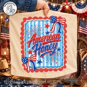 May include: A beige t-shirt featuring a patriotic design with the words "American Honey" in red and blue script. The design includes fireworks, stars, and bow accents in red, white, and blue. The shirt is held up by a person.