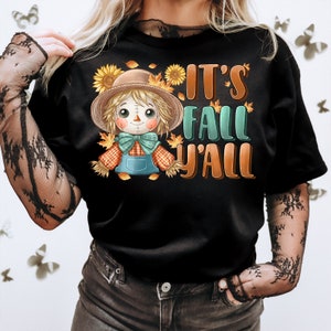 Op de afbeelding: Zwart T-shirt met een cartoon-vogelverschrikker die een bruine hoed en een geruit overhemd draagt. De tekst "It's Fall Y'all" staat op het shirt gedrukt.