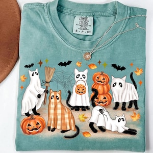 May include: A teal Comfort Colors t-shirt with a Halloween design. The graphic shows cartoon cats in ghost costumes, pumpkins, bats, and autumn leaves. A silver necklace with a pendant is also visible.